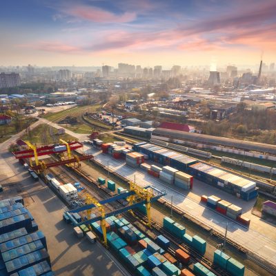 Aerial view of container loading and unloading at sunset. Top view of containers at logistics terminal in city, factory, train, railroad, sky. Business. Freight transportation containers on railway